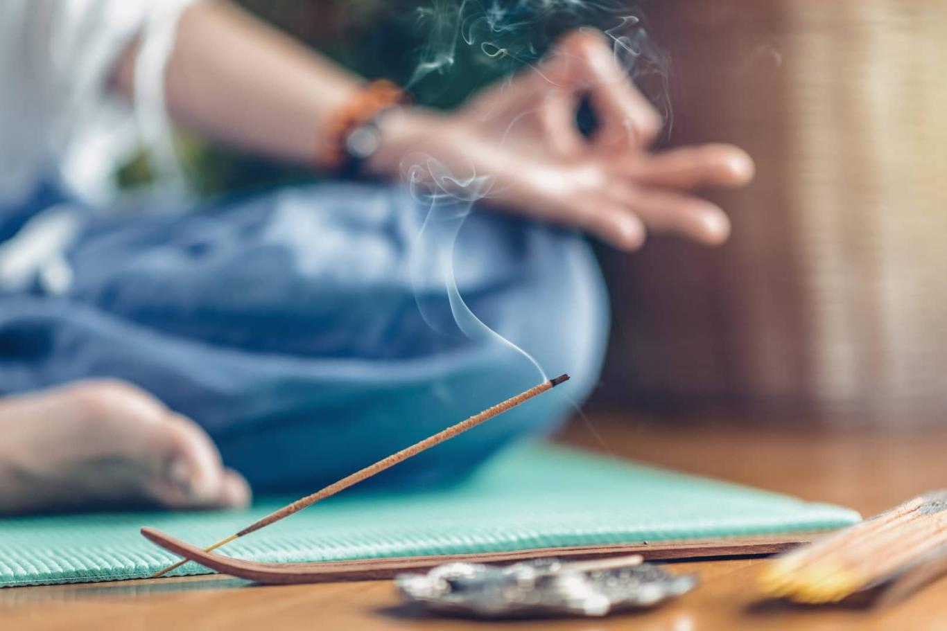 Woman meditating in lotus position on turquoise yoga mat on wooden floor. Focus on incense stick and smoke. Unrecognizable yoga practitioner in the background. Relax after Yoga training.